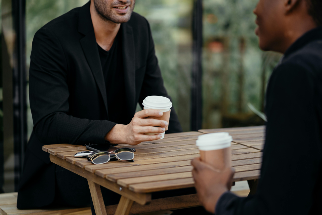 Two adults enjoying a coffee meeting outdoors, holding disposable cups on a wooden table.