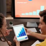 A team of colleagues discussing sales growth using a digital tablet and computer screen in an office setting.