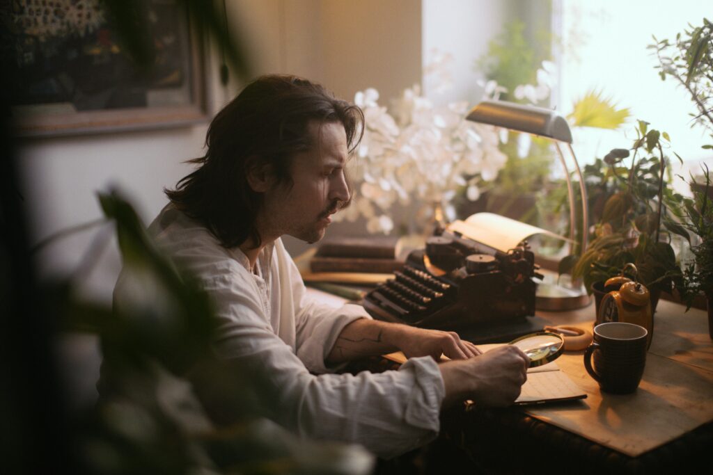 A man with long hair examines a manuscript using a magnifying glass in a cozy study.